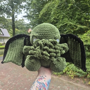 Könnte beinhalten: Grünes Häkel-Plüschtier mit schwarzen Flügeln, das einem Cthulhu-Monster ähnelt. Das Spielzeug wird von einer Hand mit einem bunten Tattoo gehalten.