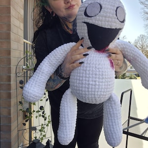 Könnte beinhalten: Ein weißes, gehäkeltes Plüschtier mit einem großen Lächeln und schwarzen Augen. Das Spielzeug hat einen roten Fleck auf der Brust. Das Spielzeug wird von einer Person mit grünen Haaren gehalten.