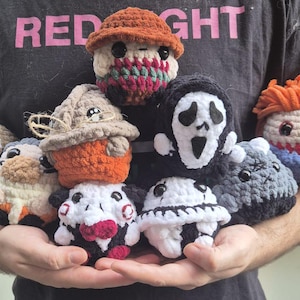 May include: A collection of crocheted horror character plushies. The plushies are in various colors, including black, white, red, orange, and blue. They are all holding different poses, and some have facial expressions. The plushies are all made of yarn and have a soft, cuddly texture.