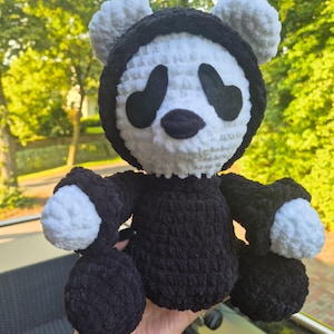 May include: A handmade crochet panda bear doll. The panda has a black body and head covering, with white accents on the face, ears, and paws. The eyes and nose are black. The doll is soft and plush, with a cuddly appearance.