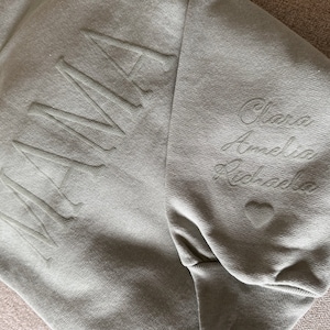 K&ouml;nnte beinhalten: Salbeigr&uuml;nes Sweatshirt mit dem Wort "MAMA" auf der Vorderseite. Der &Auml;rmel zeigt die Namen "Clara, Amelia, Richada" in Schreibschrift gestickt, zusammen mit einem kleinen Herz.