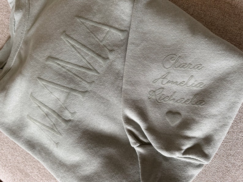 K&ouml;nnte beinhalten: Salbeigr&uuml;nes Sweatshirt mit dem Wort "MAMA" auf der Vorderseite. Der &Auml;rmel zeigt die Namen "Clara, Amelia, Richada" in Schreibschrift gestickt, zusammen mit einem kleinen Herz.