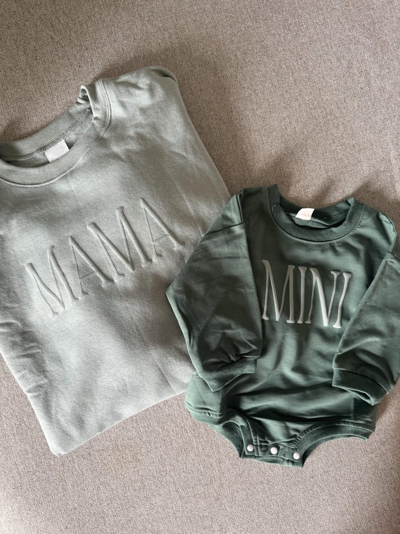 K&ouml;nnte beinhalten: Zwei passende, salbeigr&uuml;ne Sweatshirts. Eines ist ein Rundhals-Sweatshirt mit dem Wort "MAMA" auf der Vorderseite. Das andere ist ein lang&auml;rmliger Strampler mit dem Wort "MINI" auf der Vorderseite. Beide Artikel sind aus weichem Material.