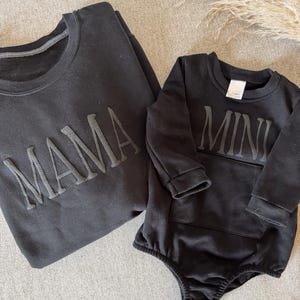 K&ouml;nnte beinhalten: Schwarzes Sweatshirt mit dem Wort "MAMA" in erhabenen Buchstaben. Daneben ein schwarzer Baby-Body mit dem Wort "MINI" in erhabenen Buchstaben. Die Artikel werden auf einer neutralen grauen Oberfl&auml;che pr&auml;sentiert.