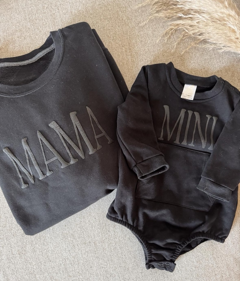 K&ouml;nnte beinhalten: Schwarzes Sweatshirt mit dem Wort "MAMA" in erhabenen Buchstaben. Daneben ein schwarzer Baby-Body mit dem Wort "MINI" in erhabenen Buchstaben. Die Artikel werden auf einer neutralen grauen Oberfl&auml;che pr&auml;sentiert.