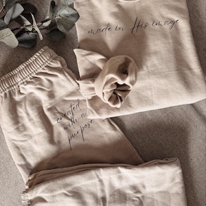 May include: A beige loungewear set featuring a sweatshirt and sweatpants. The sweatshirt has the text "made in His image" in a script font. The sweatpants have the text "created with a purpose". The set is made of a soft material.