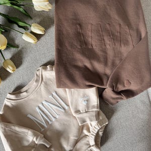 K&ouml;nnte beinhalten: Ein braunes Sweatshirt mit dem Wort "MAMA" auf der Vorderseite und ein beiger Strampler mit dem Wort "MINI" auf der Vorderseite. Beide Kleidungsst&uuml;cke haben ein Herzdetail. Die Artikel werden auf einer grauen Oberfl&auml;che pr&auml;sentiert.