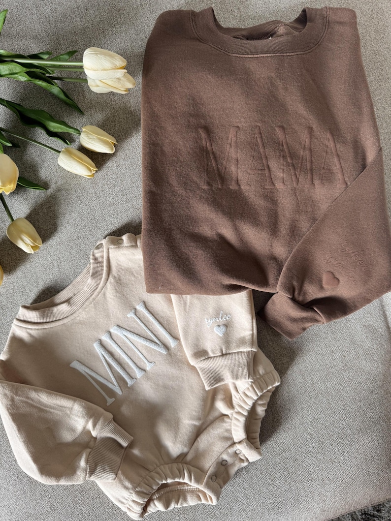 K&ouml;nnte beinhalten: Ein braunes Sweatshirt mit dem Wort "MAMA" auf der Vorderseite und ein beiger Strampler mit dem Wort "MINI" auf der Vorderseite. Beide Kleidungsst&uuml;cke haben ein Herzdetail. Die Artikel werden auf einer grauen Oberfl&auml;che pr&auml;sentiert.