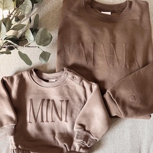 Może przedstawiać: Brązowa bluza z napisem "MAMA" i brązowe body dla dziecka z wytłoczonym napisem "MINI" z przodu. Body ma długie rękawy i zatrzaski na ramieniu. Bluza ma długie rękawy.