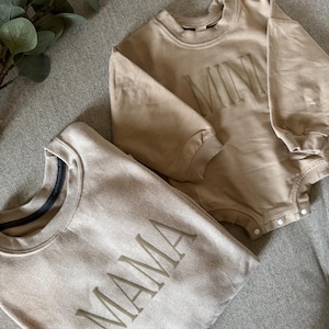 K&ouml;nnte beinhalten: Zwei beige Sweatshirts und ein Strampler. Auf einem Sweatshirt ist das Wort "MAMA" auf der Vorderseite gestickt. Der Strampler hat das Wort "MINI" auf der Vorderseite gestickt. Die Kleidungsst&uuml;cke sind aus weichem Material.