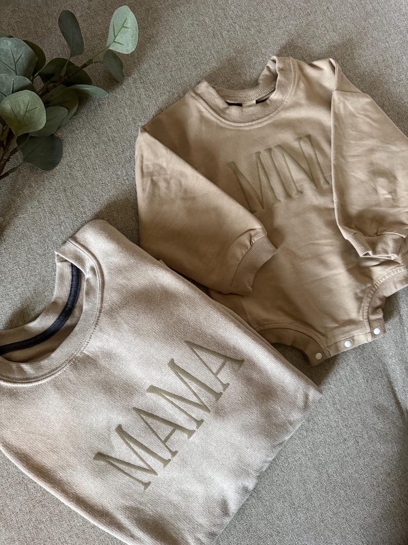 K&ouml;nnte beinhalten: Zwei beige Sweatshirts und ein Strampler. Auf einem Sweatshirt ist das Wort "MAMA" auf der Vorderseite gestickt. Der Strampler hat das Wort "MINI" auf der Vorderseite gestickt. Die Kleidungsst&uuml;cke sind aus weichem Material.