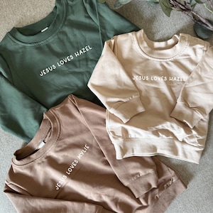 May include: Three crewneck sweatshirts in olive green, tan, and brown. Each sweatshirt has the phrase "JESUS LOVES HAZEL" or "JESUS LOVES MYLIE" printed in white. The sweatshirts have ribbed cuffs and a ribbed hem.