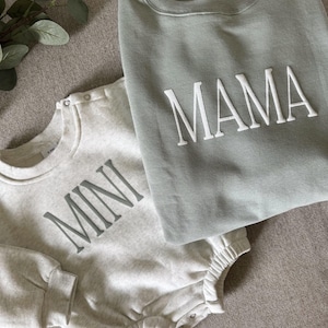 K&ouml;nnte beinhalten: Ein hellgrauer Baby-Strampler mit dem Wort "MINI" in grauen Buchstaben und ein salbeigr&uuml;nes Sweatshirt mit dem Wort "MAMA" in wei&szlig;en Buchstaben. Die Kleidungsst&uuml;cke sind aus einem weichen Material und auf einer neutralen Oberfl&auml;che gefaltet.