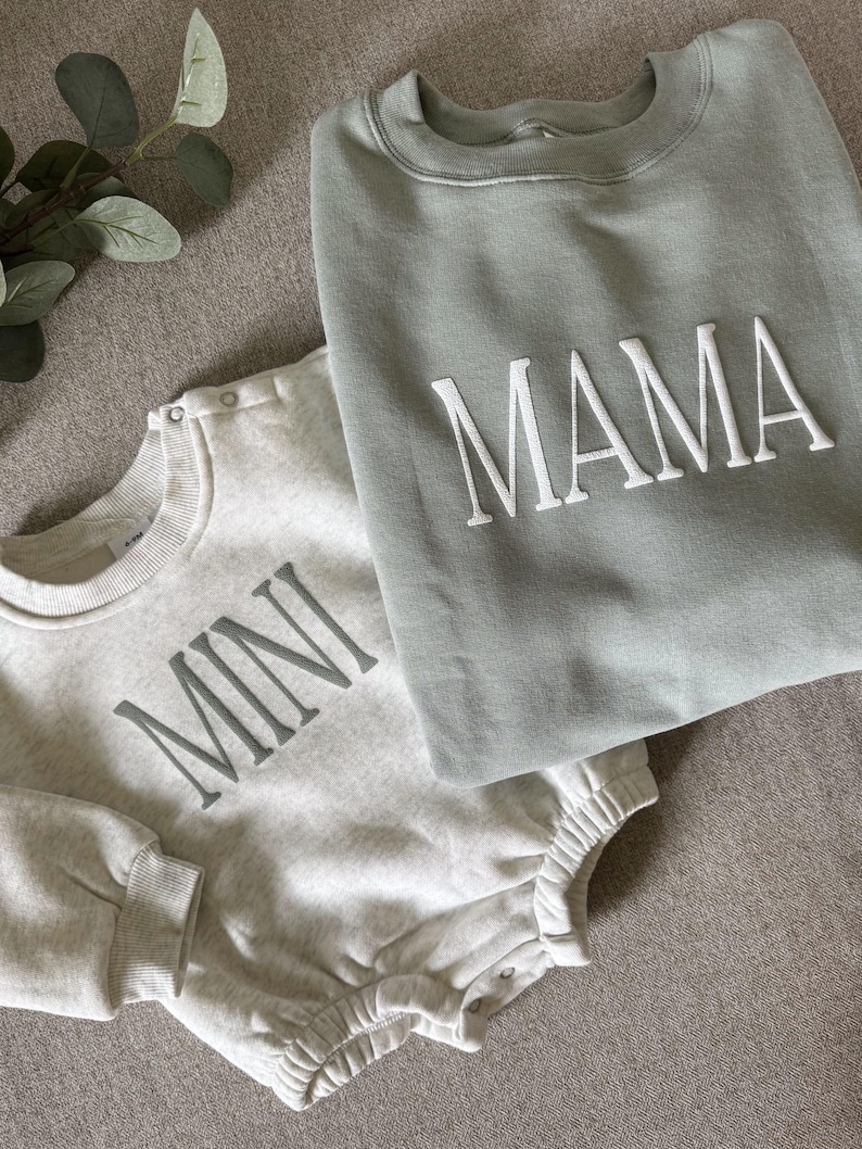 K&ouml;nnte beinhalten: Ein hellgrauer Baby-Strampler mit dem Wort "MINI" in grauen Buchstaben und ein salbeigr&uuml;nes Sweatshirt mit dem Wort "MAMA" in wei&szlig;en Buchstaben. Die Kleidungsst&uuml;cke sind aus einem weichen Material und auf einer neutralen Oberfl&auml;che gefaltet.