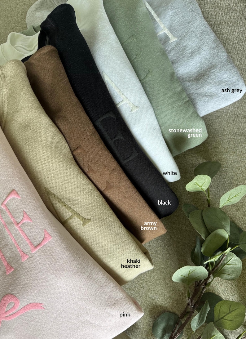 K&ouml;nnte beinhalten: Gefaltete Kapuzenpullover in verschiedenen Farben, darunter Rosa, Khaki-Melange, Armeebraun, Schwarz, Wei&szlig;, Stone Washed Gr&uuml;n und Aschgrau. Jeder Hoodie hat ein gesticktes Buchstabendesign. Draufsicht.