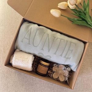 Könnte beinhalten: Geöffnete Geschenkbox aus Pappe mit einer hellblauen Decke mit dem Wort "AUNTIE", einem kleinen weißen Handtuch, einer Kerze im Glas und einer Holzblume. Gelbe Tulpen liegen auf der Box. Die Box ist mit Papier gefüllt.