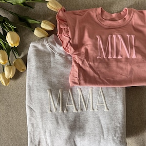 K&ouml;nnte beinhalten: Ein hellgraues Sweatshirt mit dem Wort "MAMA" in Wei&szlig; gestickt, kombiniert mit einem rosa T-Shirt mit gesticktem "MINI". Das T-Shirt hat R&uuml;schen&auml;rmel. Gelbe Tulpen sind im Hintergrund zu sehen.
