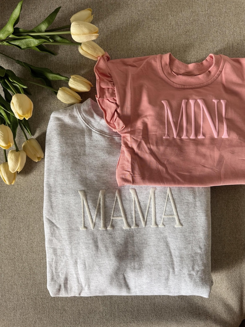 K&ouml;nnte beinhalten: Ein hellgraues Sweatshirt mit dem Wort "MAMA" in Wei&szlig; gestickt, kombiniert mit einem rosa T-Shirt mit gesticktem "MINI". Das T-Shirt hat R&uuml;schen&auml;rmel. Gelbe Tulpen sind im Hintergrund zu sehen.