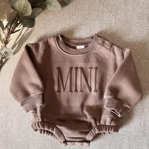 K&ouml;nnte beinhalten: Ein brauner Baby-Strampler mit dem Wort &bdquo;MINI&ldquo; auf der Vorderseite. Der lang&auml;rmlige Strampler hat einen Rundhalsausschnitt und Druckkn&ouml;pfe an der Schulter. Der untere Teil des Stramplers ist mit einem Gummizug gerafft. Der Strampler ist aus weichem Material.
