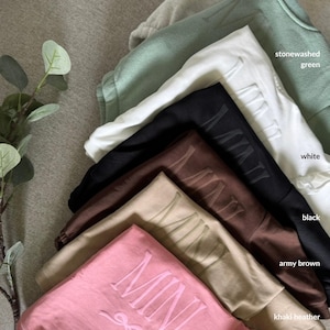 K&ouml;nnte beinhalten: Eine Auswahl an Sweatshirts in verschiedenen Farben, darunter Aschgrau, Stone Washed Gr&uuml;n, Wei&szlig;, Schwarz, Armeeblau, Khaki Heather und Pink. Jedes Sweatshirt tr&auml;gt das Wort "MINI" in einer passenden Farbe.