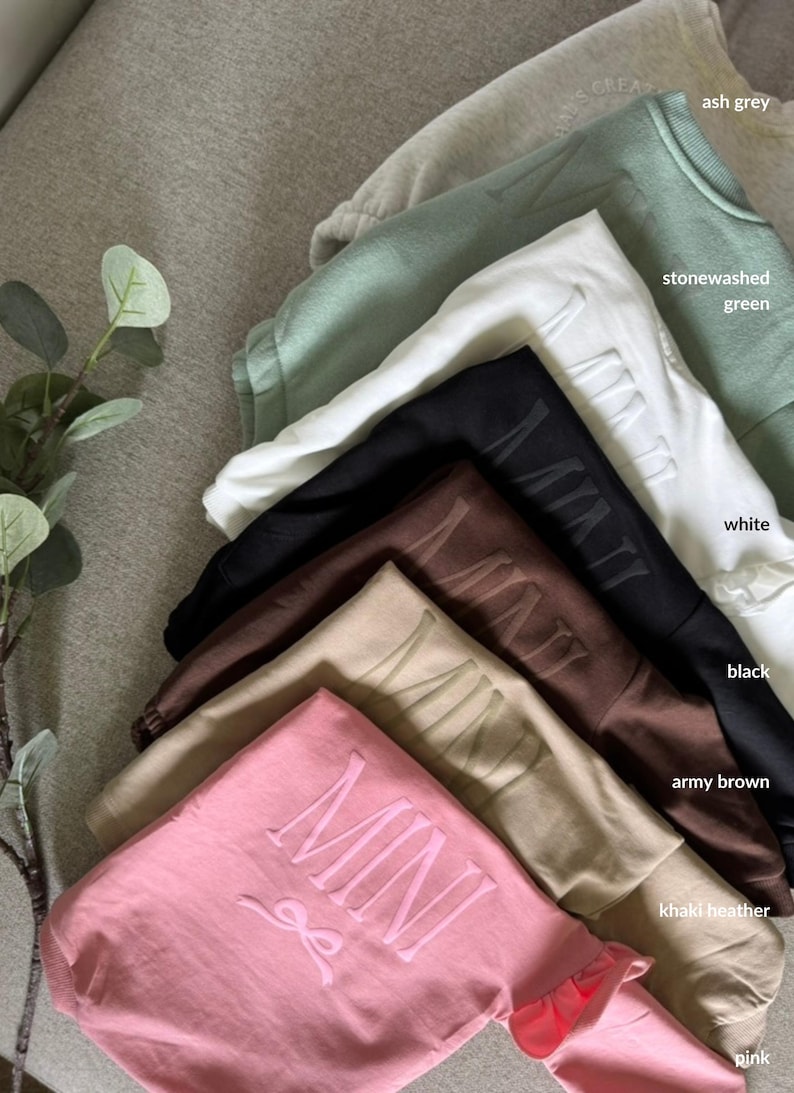 K&ouml;nnte beinhalten: Eine Auswahl an Sweatshirts in verschiedenen Farben, darunter Aschgrau, Stone Washed Gr&uuml;n, Wei&szlig;, Schwarz, Armeeblau, Khaki Heather und Pink. Jedes Sweatshirt tr&auml;gt das Wort "MINI" in einer passenden Farbe.