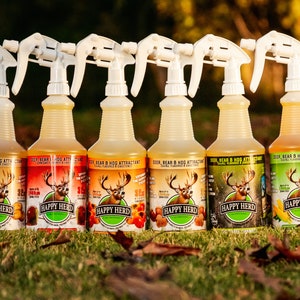 May include: A row of seven plastic spray bottles filled with a yellow liquid, labeled "Deer, Bear & Hog Attractant" with the "Happy Herd" logo. The bottles are on a grassy surface with fallen leaves.