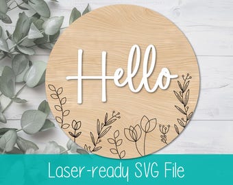 May include: A round wooden sign with the word "Hello" in white lettering and a black floral design around the bottom edge. The sign is laser-ready for crafting.