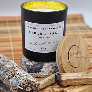 May include: Black soy candle with a gold lid and a white label that reads "Goodnight Moon Candle Co. Cedar & Sage Soy Candle Made with Magic 10oz". The candle is sitting on a woven bamboo mat with a bundle of sage, palo santo sticks, and a wooden lid with the Goodnight Moon Candle Co. logo.