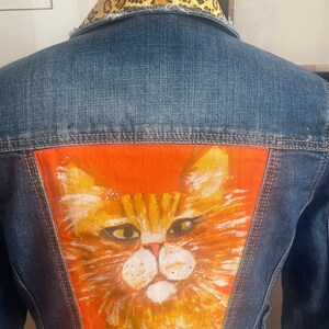 May include: A blue denim jacket with a leopard print collar and a patch of a painted orange cat on the back.