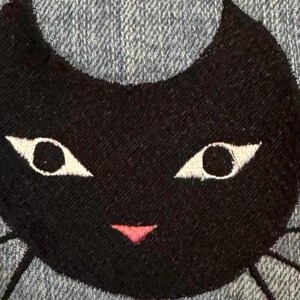 May include: A black cat face embroidered on a blue denim fabric. The cat has white eyes and a pink mouth.