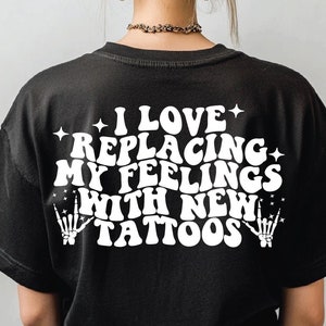 Może przedstawiać: Czarny T-shirt z białym tekstem, który brzmi "Uwielbiam zastępować swoje uczucia nowymi tatuażami". Tekst jest w retro stylu czcionki z gwiazdami i szkieletowymi dłońmi po obu stronach.