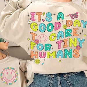 May include: A cream-colored sweatshirt with a colorful graphic print that says "It's a good day to care for tiny humans". The graphic features a smiley face, flowers, stars, and a butterfly.
