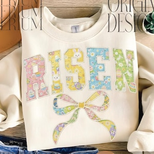 May include: Cream-colored sweatshirt with the word "RISEN" in pastel floral and bunny-themed letters. A large bow with pastel floral patterns is below the word. The sweatshirt is paired with denim shorts and a brown leather sandal.