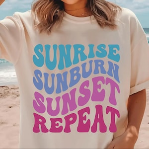 Peut inclure: T-shirt blanc avec un imprim&eacute; graphique du texte "Sunrise Sunburn Sunset Repeat" en bleu, violet et rose.