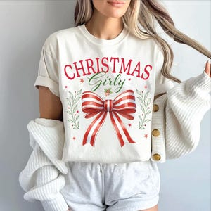 Puede incluir: Camiseta blanca con el texto "Christmas Girly" en letras rojas y verdes. Un lazo rojo y blanco a rayas con hojas verdes est&aacute; centrado en la camiseta.
