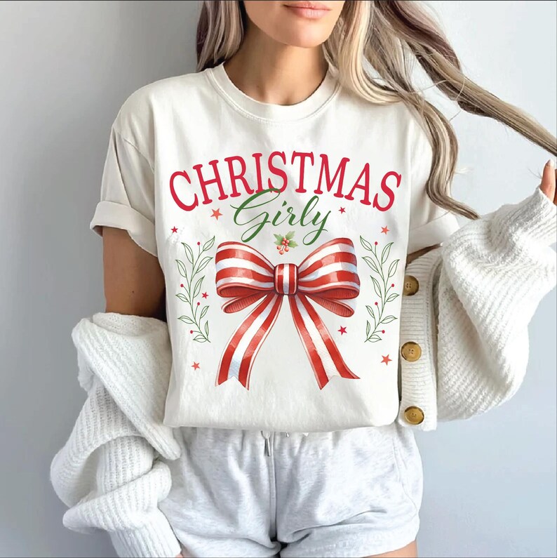 Puede incluir: Camiseta blanca con el texto "Christmas Girly" en letras rojas y verdes. Un lazo rojo y blanco a rayas con hojas verdes est&aacute; centrado en la camiseta.