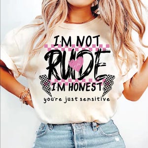 May include: White t-shirt with black text that reads "I'm not rude I'm honest you're just sensitive". The text is surrounded by a pink and black checkered pattern and a pink heart.
