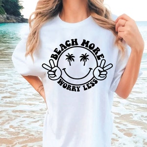 以下が含まれることがあります： 白いTシャツに、黒いスマイリーフェイスのグラフィックが描かれており、目はヤシの木、テキストは「ビーチモアワーリーレス」です。