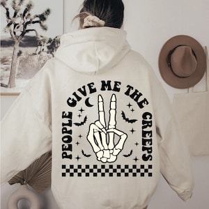 Op de afbeelding: Een cr&egrave;mekleurige hoodie met een zwarte afbeelding op de rug. De afbeelding toont een skelethand die een vredesteken maakt met de tekst "People Give Me The Creeps" eromheen.