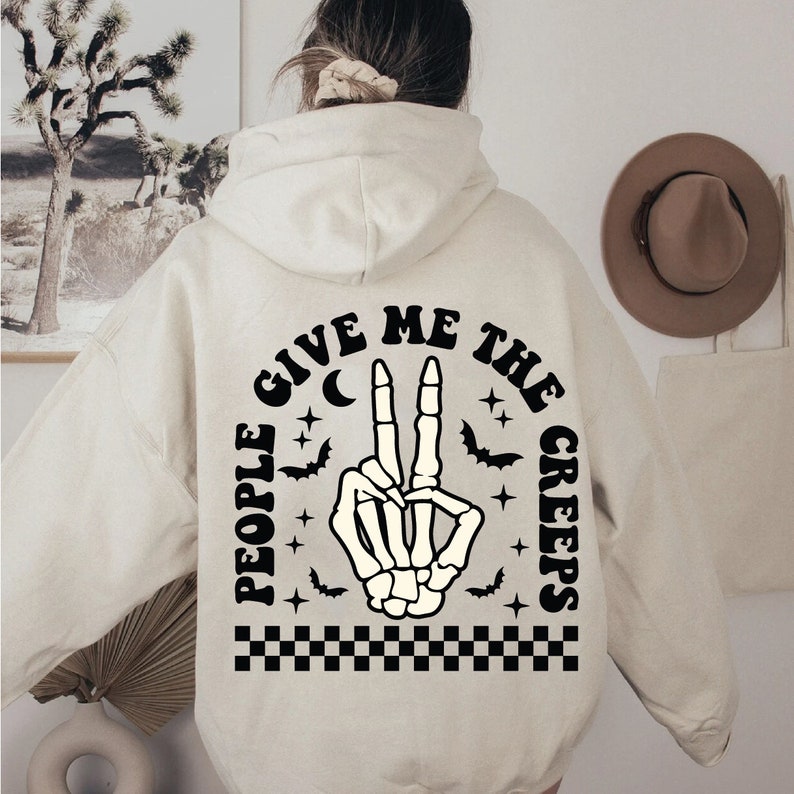 Op de afbeelding: Een cr&egrave;mekleurige hoodie met een zwarte afbeelding op de rug. De afbeelding toont een skelethand die een vredesteken maakt met de tekst "People Give Me The Creeps" eromheen.