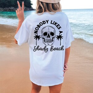 以下が含まれることがあります： 黒い頭蓋骨とヤシの木のグラフィックがプリントされた白いTシャツ。グラフィックの下に「Nobody Likes a Shady Beach」というテキストがプリントされています。