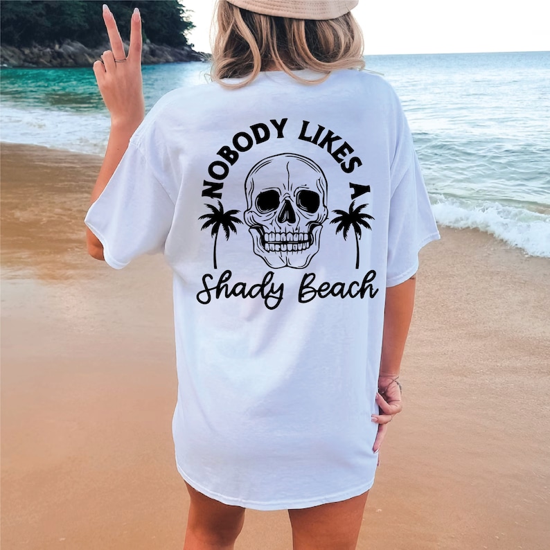 以下が含まれることがあります： 黒い頭蓋骨とヤシの木のグラフィックがプリントされた白いTシャツ。グラフィックの下に「Nobody Likes a Shady Beach」というテキストがプリントされています。