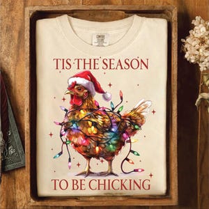May include: A white t-shirt with a Christmas-themed design featuring a chicken wrapped in colorful Christmas lights. The text "Tis the season to be chicking" is printed in red on the shirt.