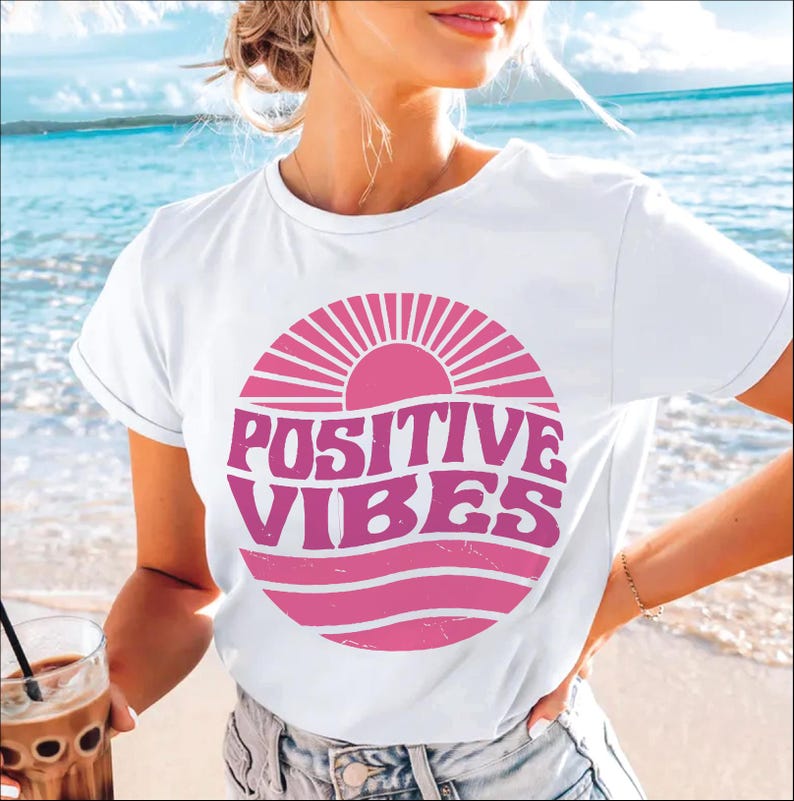 Peut inclure: T-shirt blanc avec un graphique rose d'un soleil et des vagues avec le texte "POSITIVE VIBES" dans une police r&eacute;tro.