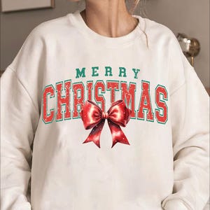 Puede incluir: Sudadera blanca con un gr&aacute;fico vintage rojo y verde que dice "Merry Christmas" con un lazo rojo.