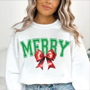Puede incluir: Sudadera blanca con un gr&aacute;fico verde "MERRY" y un lazo rojo.