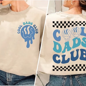 May include: A beige sweatshirt with a blue and green "Cool Dads Club" graphic on the front and a blue and green "Cool Dads Club" graphic with a black and white chequered pattern on the back.