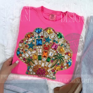 Puede incluir: Una camiseta rosa intenso con un gran diseño de concha ornamentada. La concha está adornada con gemas de colores, incluyendo azul, verde, amarillo y rojo, y presenta palmeras y estrellas de mar. La camiseta es de la marca Comfort Colors.