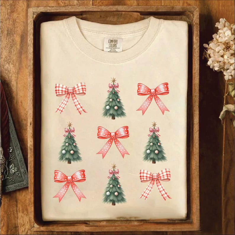 Puede incluir: Una camiseta blanca con un lazo a cuadros rojo y blanco y un &aacute;rbol de Navidad verde con adornos blancos que se repiten en todo el dise&ntilde;o. El texto "COMFY COLORS" es visible en la camiseta.