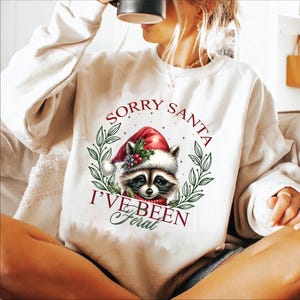 May include: White sweatshirt with a Christmas-themed raccoon wearing a Santa hat and the text "Sorry Santa I've Been Feral".